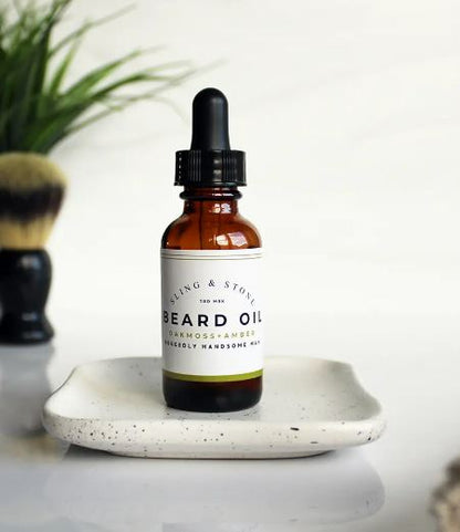 A bottle of beard oil placed on a ceramic tray with grooming accessories in the background, suggesting a men's grooming routine.
