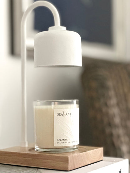 A scented candle named "sea love atlantic orange nectar ne004" rests on a wooden coaster, with a soft glow emanating from beneath a white modern-style table lamp, creating an atmosphere of warmth and tranquility.